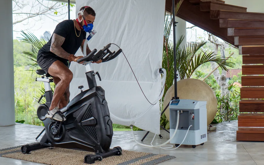 A person cycling indoors while breathing through a mask connected to an oxygen concentrator.